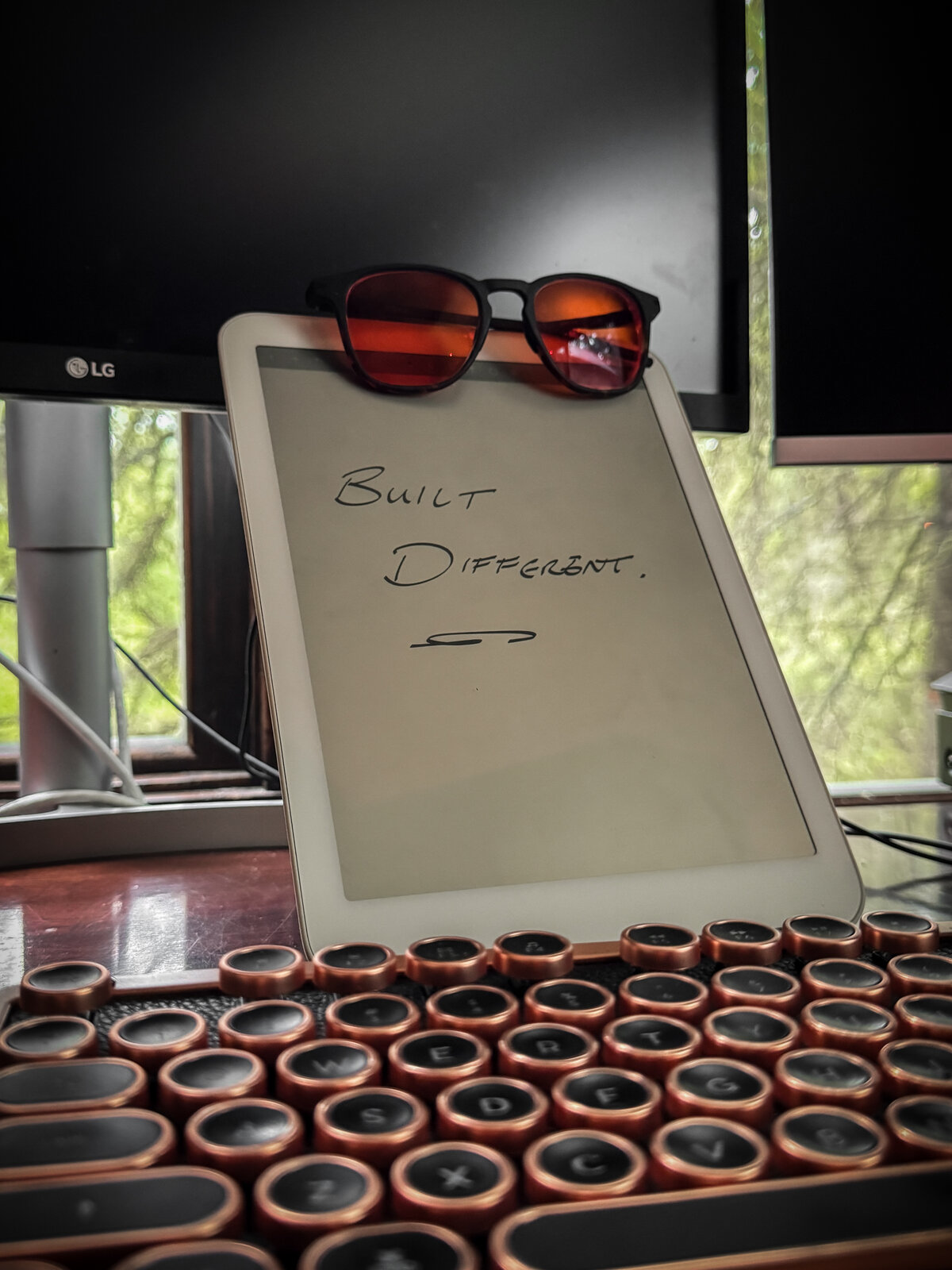 Handwritten 'Built different' on an e-ink tablet at the desk, with vintage keyboard and woods through the window